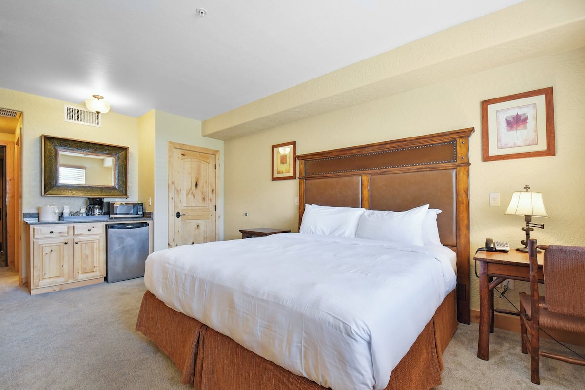 A hotel room with a large bed, a wooden headboard, a desk with a chair, a small table with a lamp, a microwave, a coffee maker, and a refrigerator.