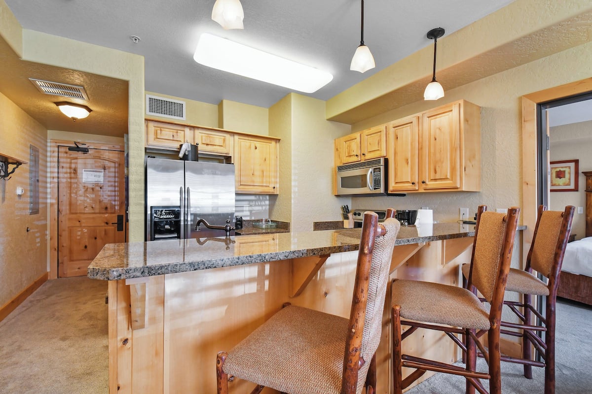 A kitchen with wooden cabinets, a stainless steel refrigerator, a microwave, and a granite countertop. There are two wicker chairs at the island.
