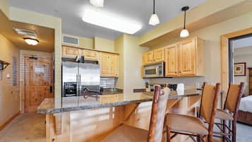A kitchen with wooden cabinets, a stainless steel refrigerator, a microwave, and a granite countertop. There are two wicker chairs at the island.