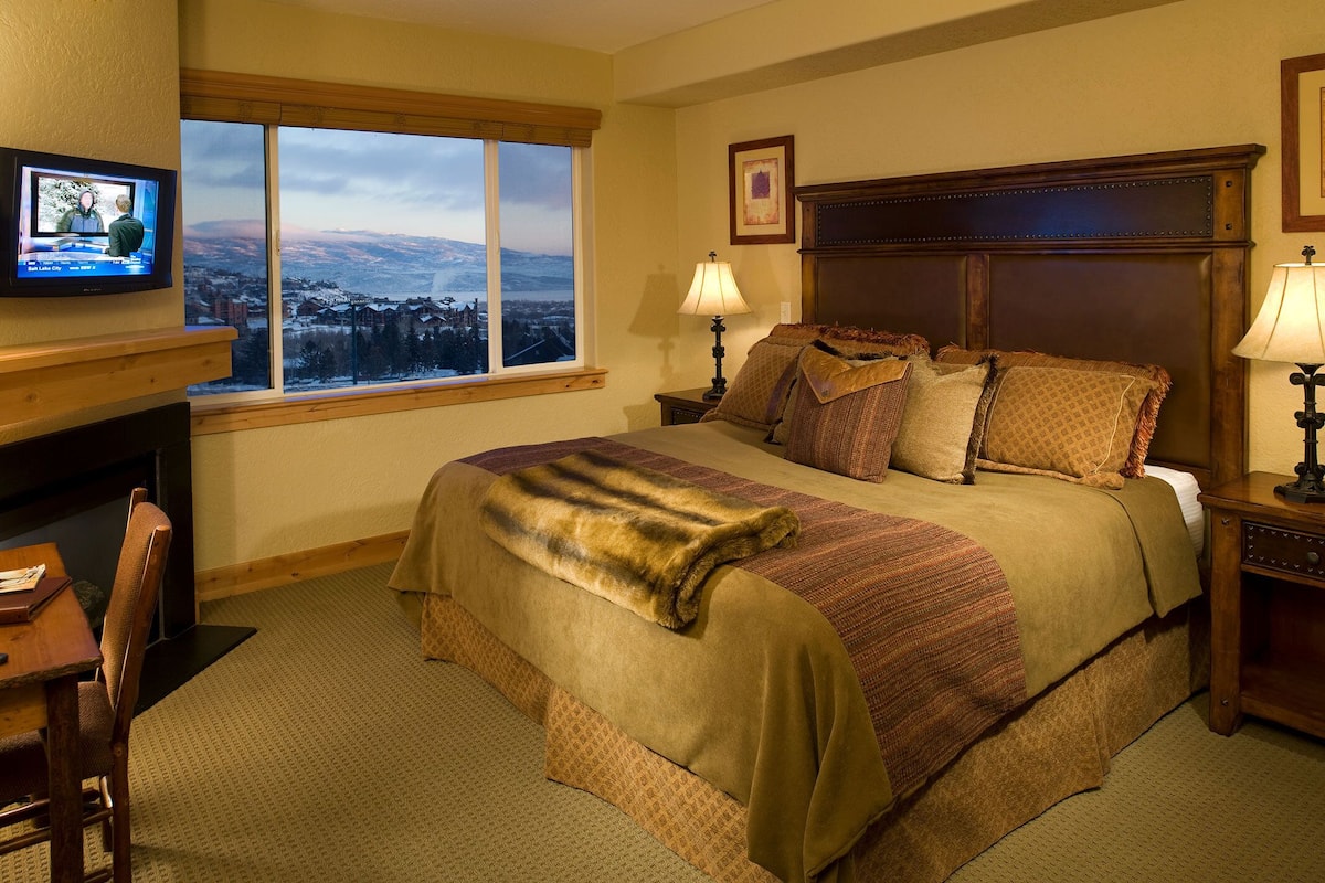 A hotel room with a large bed, two bedside lamps, a television on a desk, and a window with a view of a snowy landscape.