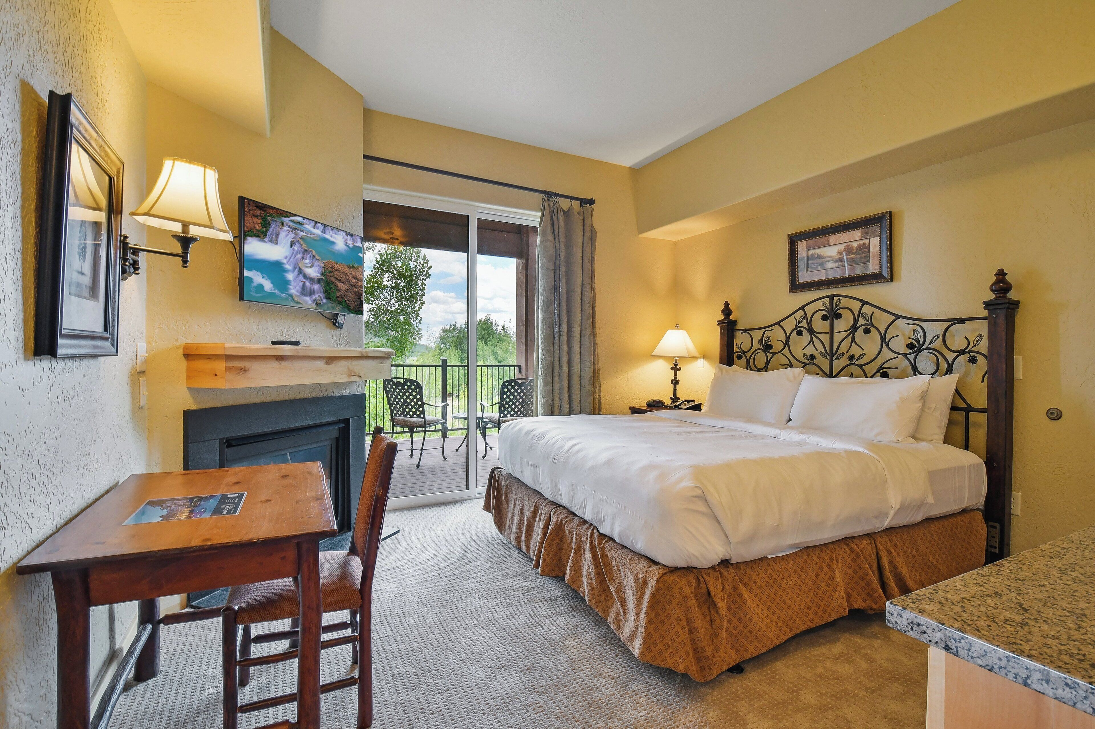A hotel room with a bed, a desk, a chair, a fireplace, a television, and a balcony with chairs.