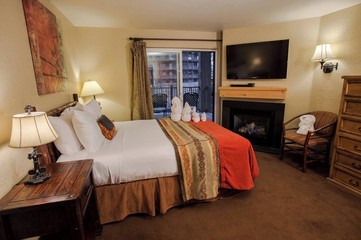 A hotel room with a bed, a TV, a fireplace, and a balcony with chairs.