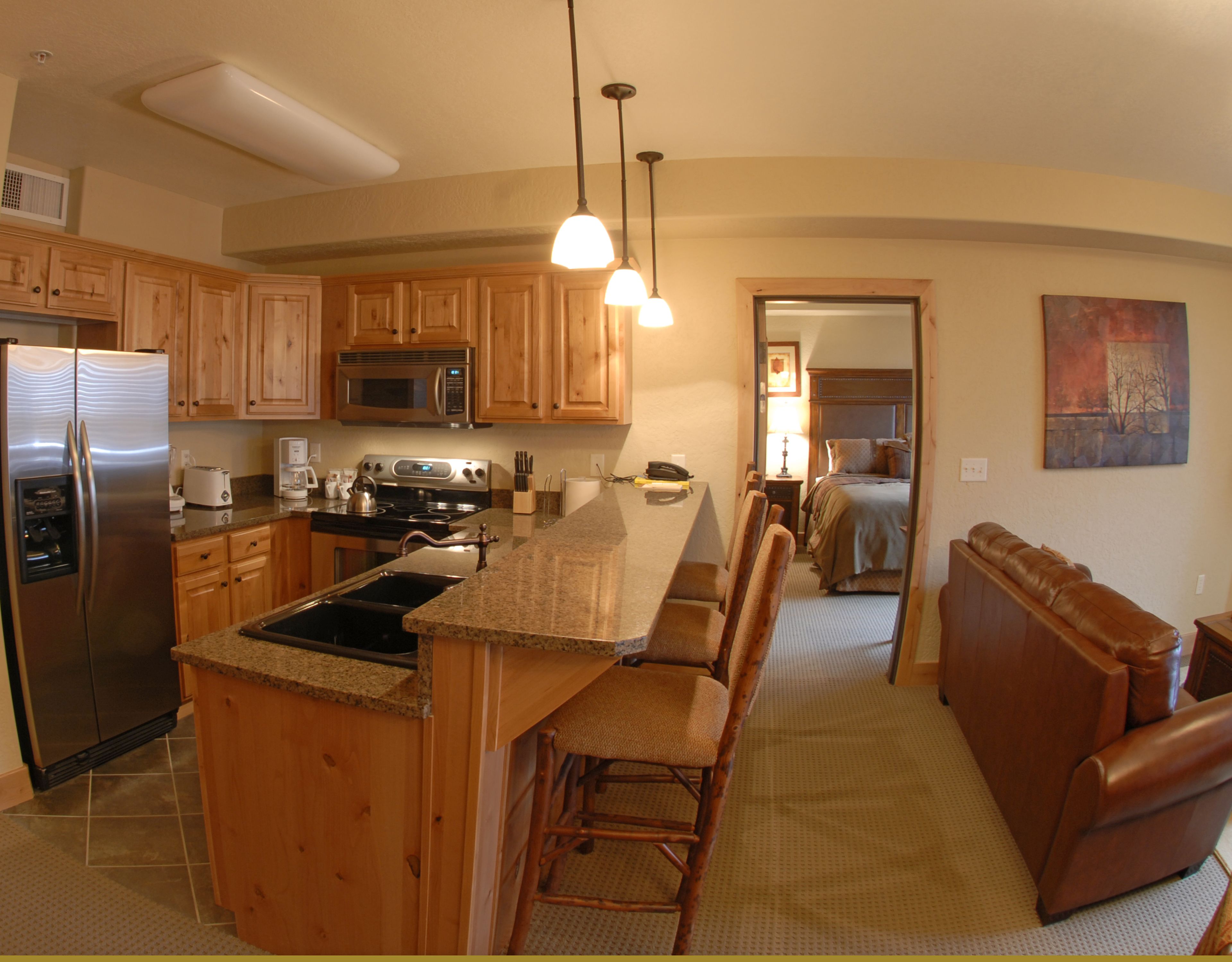 A kitchen with wooden cabinets, a stainless steel refrigerator, a microwave, and a stove. There is a breakfast bar with chairs and a brown leather couch.