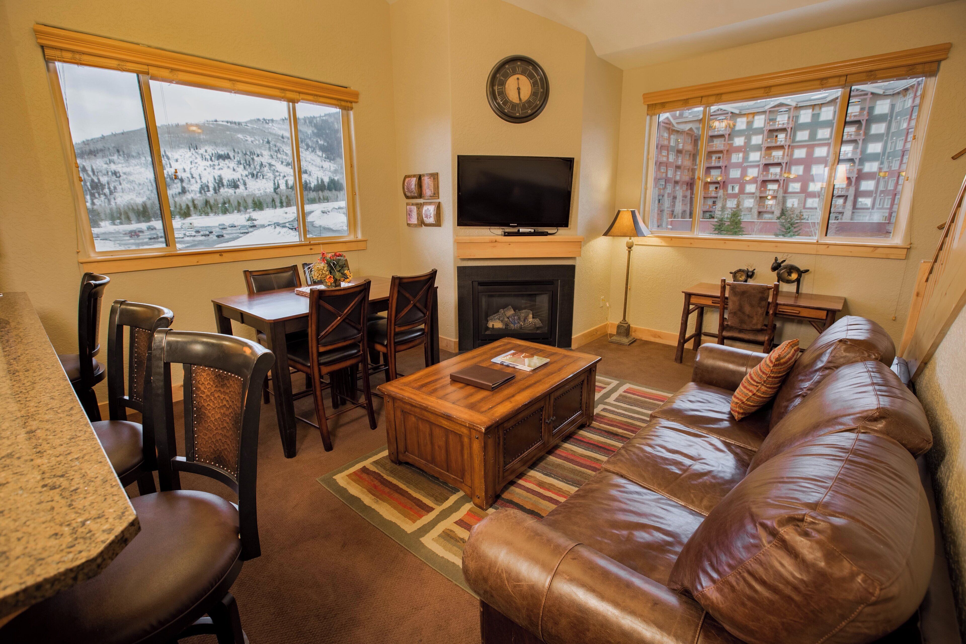 A cozy living room with a fireplace, a television, and a large window with a view of snow-covered mountains.