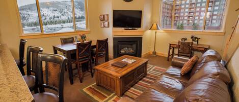A cozy living room with a fireplace, a television, and a large window with a view of snow-covered mountains.