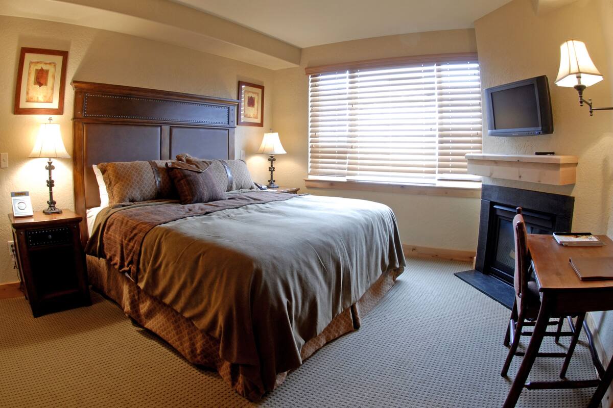 A bedroom with a large bed, two bedside lamps, a television above a fireplace, and a desk with a chair.