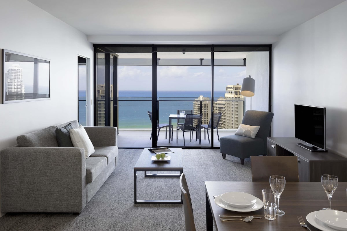 A modern living room with a sofa, a dining table set for two, a TV, and a balcony with a view of the ocean and buildings.