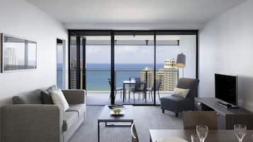 A modern living room with a sofa, a dining table set for two, a TV, and a balcony with a view of the ocean and buildings.