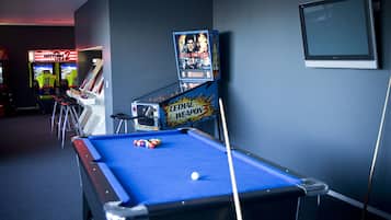 A pool table with a blue felt surface, a pool cue, and a white cue ball. There are arcade machines, including 'Daytona USA' and 'Lethal Weapon 3'. A television is mounted on the wall.