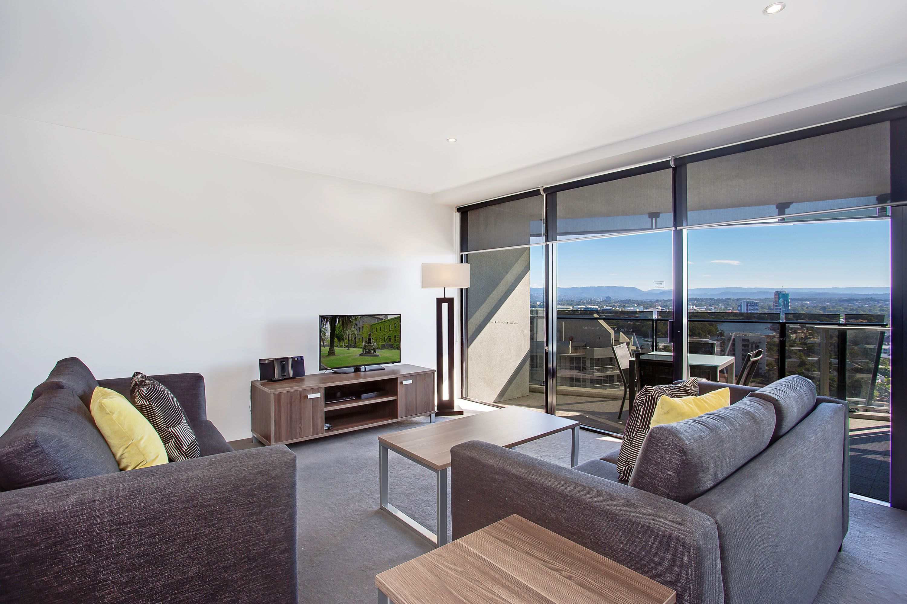 A modern living room with a large flat-screen TV, a sofa set, a coffee table, and a view of the city through floor-to-ceiling windows.