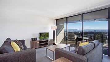 A modern living room with a large flat-screen TV, a sofa set, a coffee table, and a view of the city through floor-to-ceiling windows.