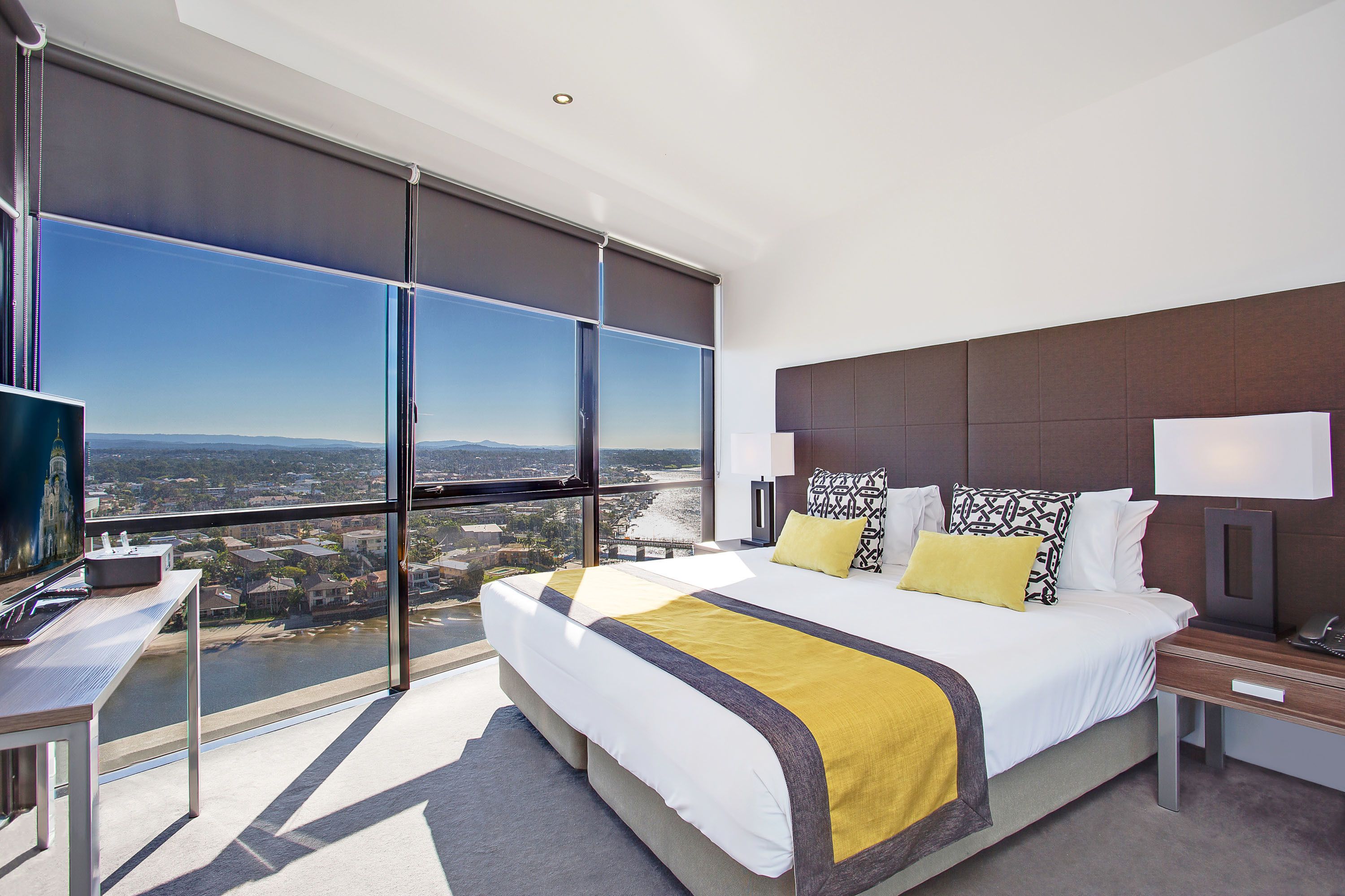 A modern hotel room with a large bed, a desk, and a view of a cityscape through floor-to-ceiling windows.