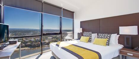 A modern hotel room with a large bed, a desk, and a view of a cityscape through floor-to-ceiling windows.