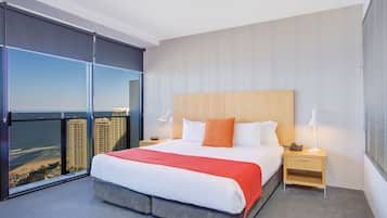 A modern hotel room with a large bed, a nightstand, a lamp, and a view of the beach and buildings.