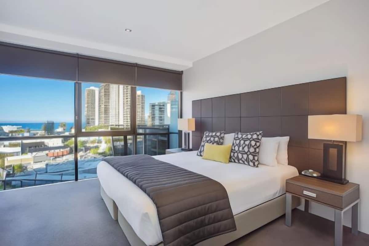 A modern hotel room with a large bed, a nightstand, a lamp, and a view of the cityscape through floor-to-ceiling windows.