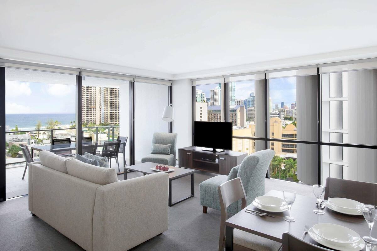A modern living room with a sofa, chairs, a dining table, and a TV. Large windows offer a city view.
