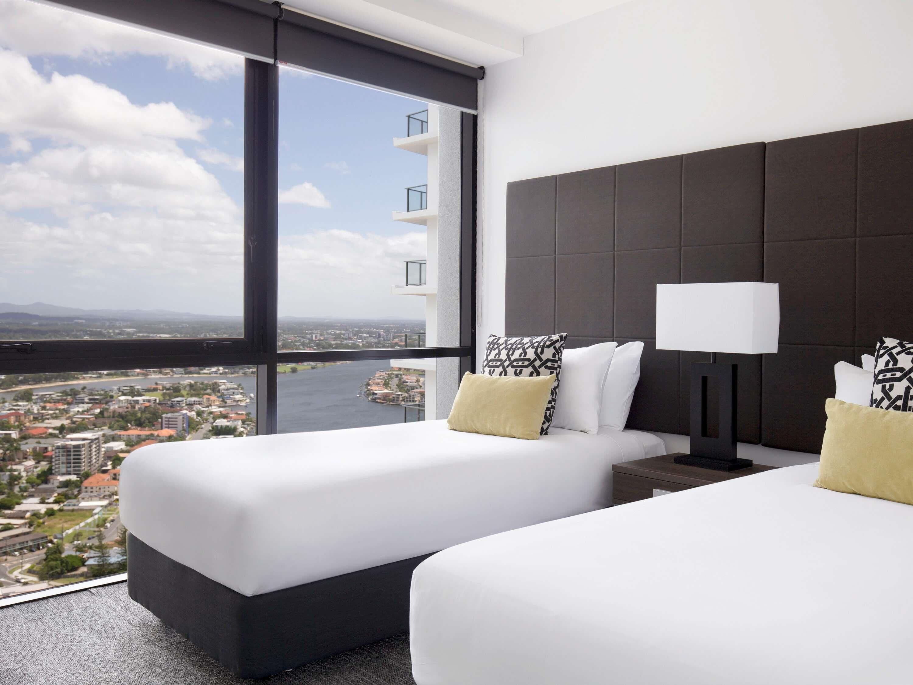 A hotel room with two beds, a large window with a city view, a dark headboard, and a bedside table with a lamp.