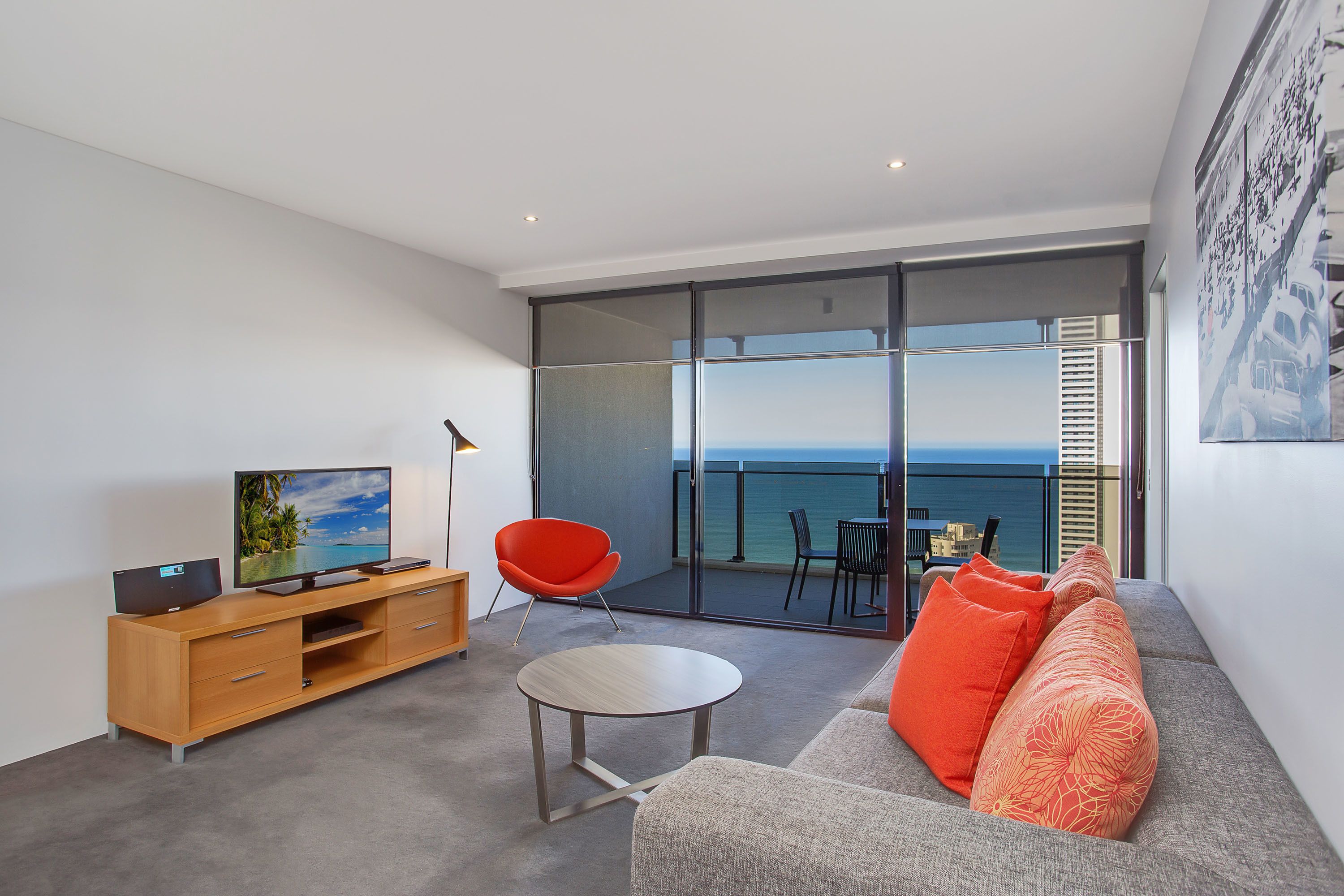 A modern living room with a sofa, a red chair, a TV, and a balcony with a view of the sea.