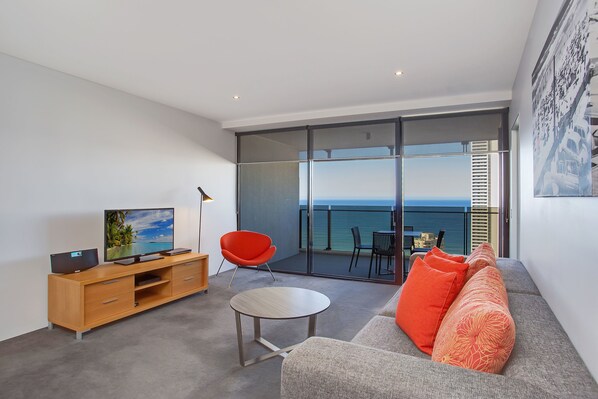 A modern living room with a sofa, a red chair, a TV, and a balcony with a view of the sea.