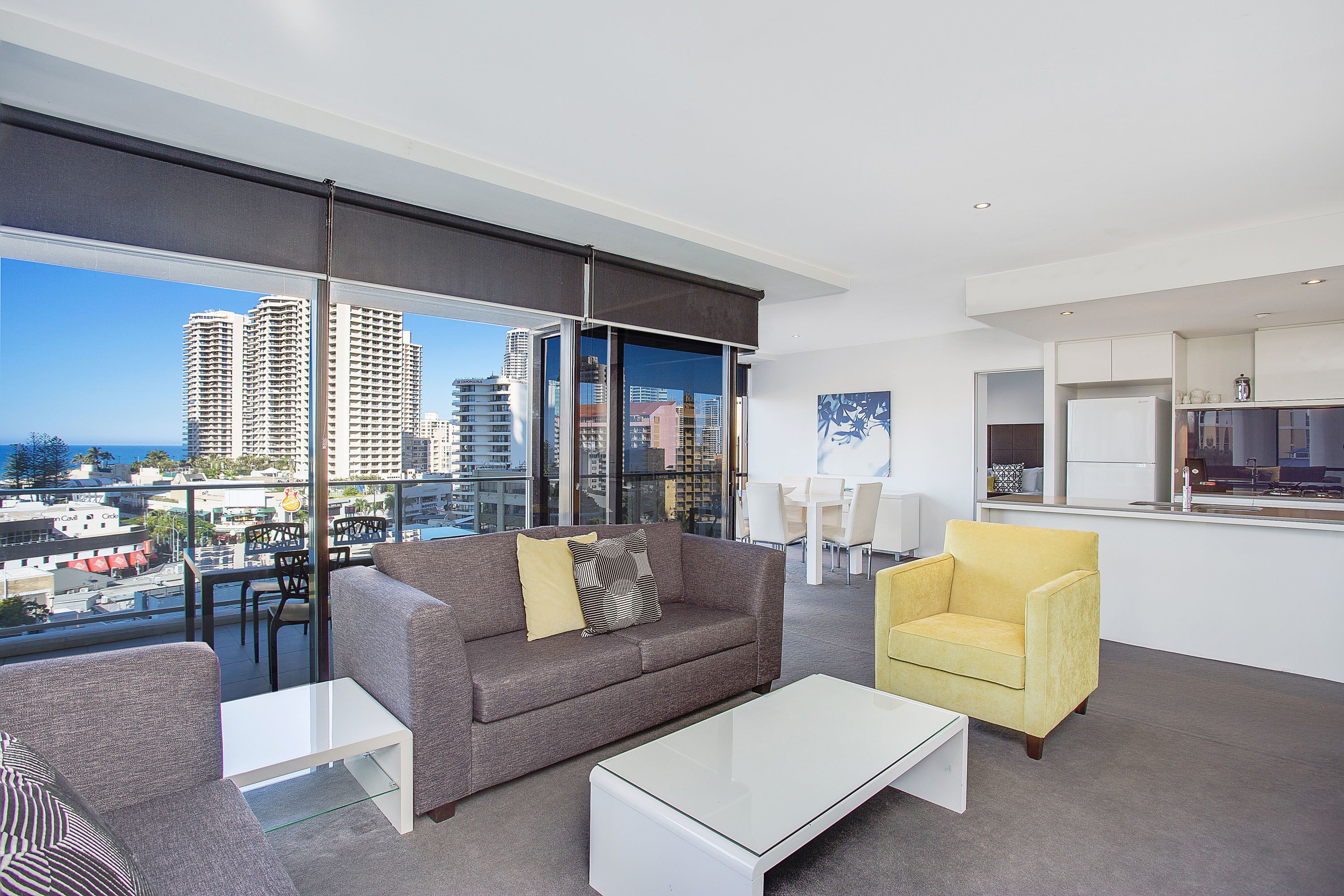 A modern living room with a grey sofa, a yellow armchair, and a white coffee table. There is a large window with a city view.