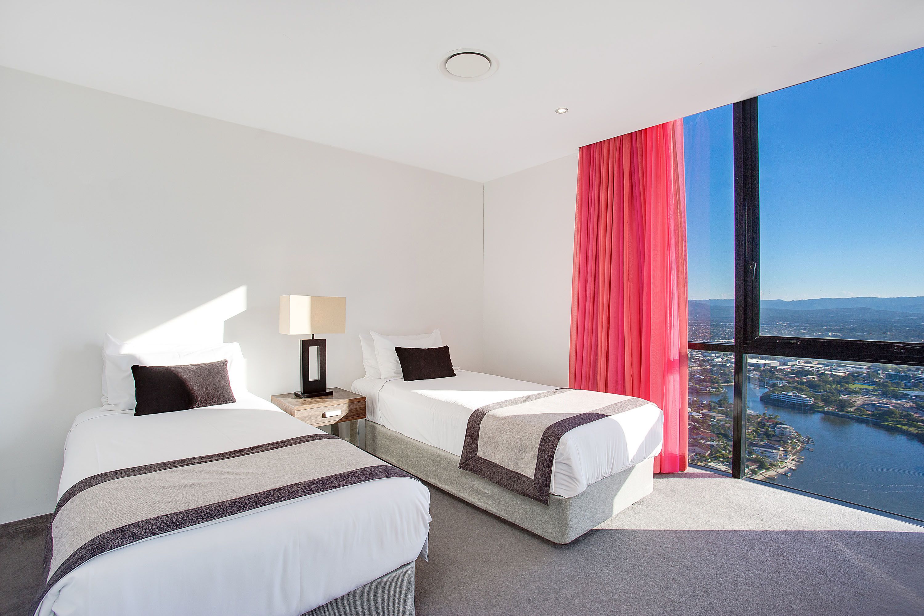 Habitación de hotel con dos camas, un ventanal grande con vistas a la ciudad y al agua, y una cortina roja.
