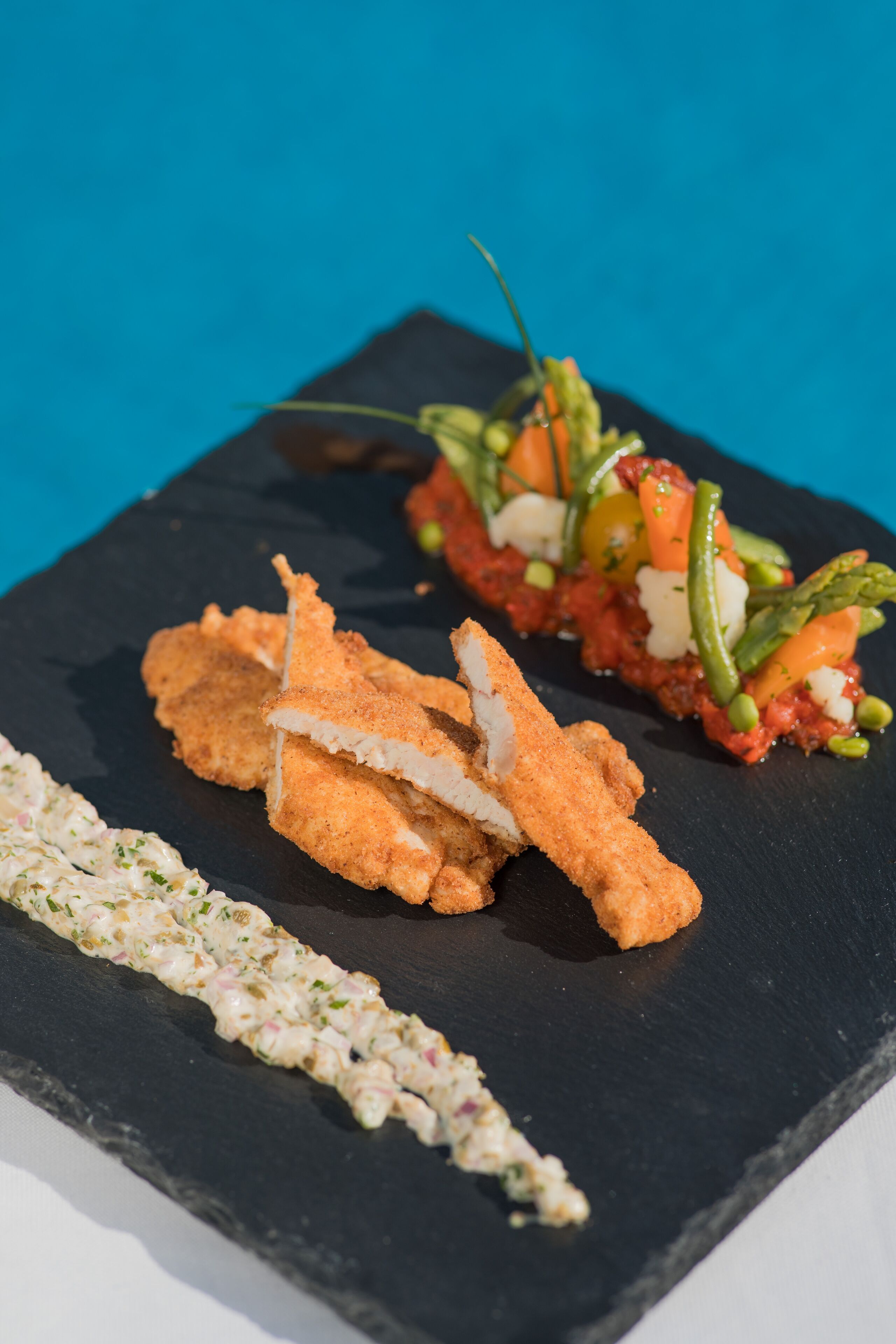 A black slate serving board with three food items: breaded fish, a vegetable and cheese salad, and a creamy spread.