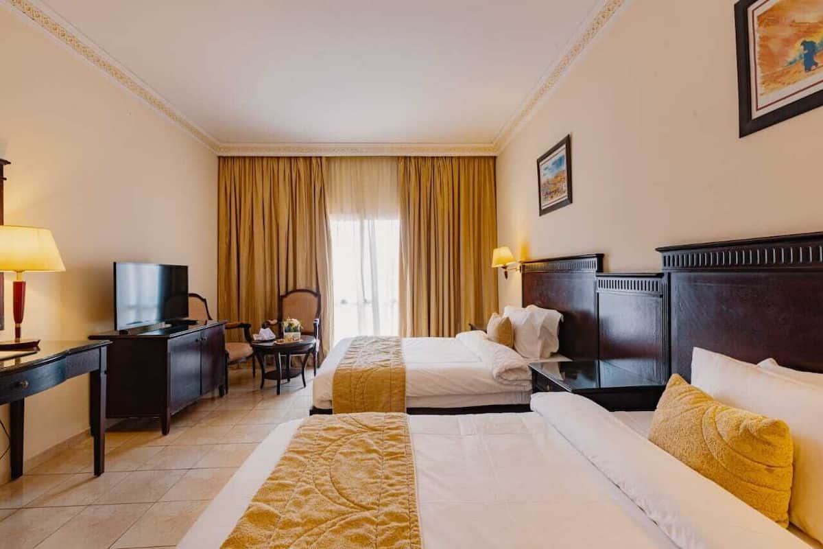A hotel room with two beds, a desk, a chair, a TV, and a large window with curtains.