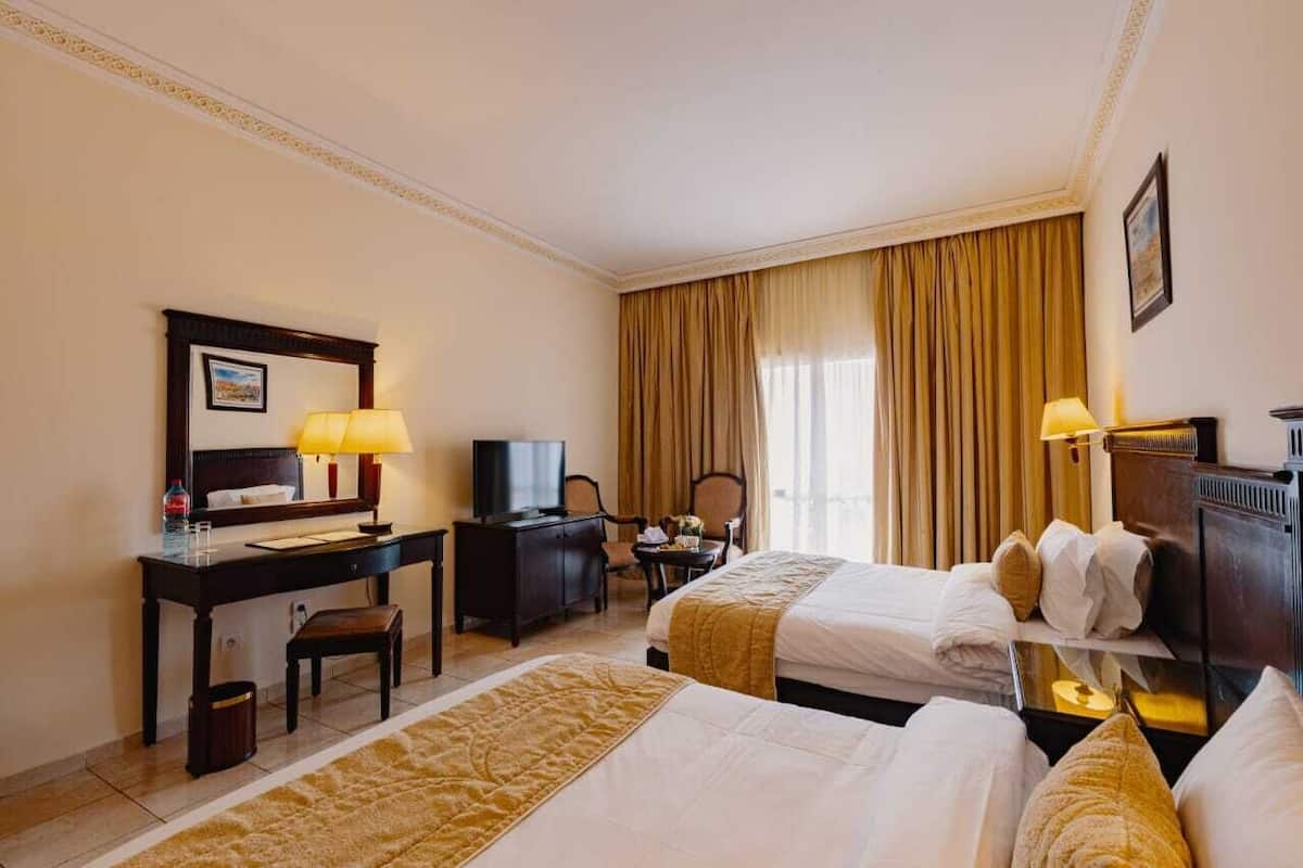 A hotel room with two beds, a desk with a computer and lamp, a chair, a TV, and a large window with curtains.
