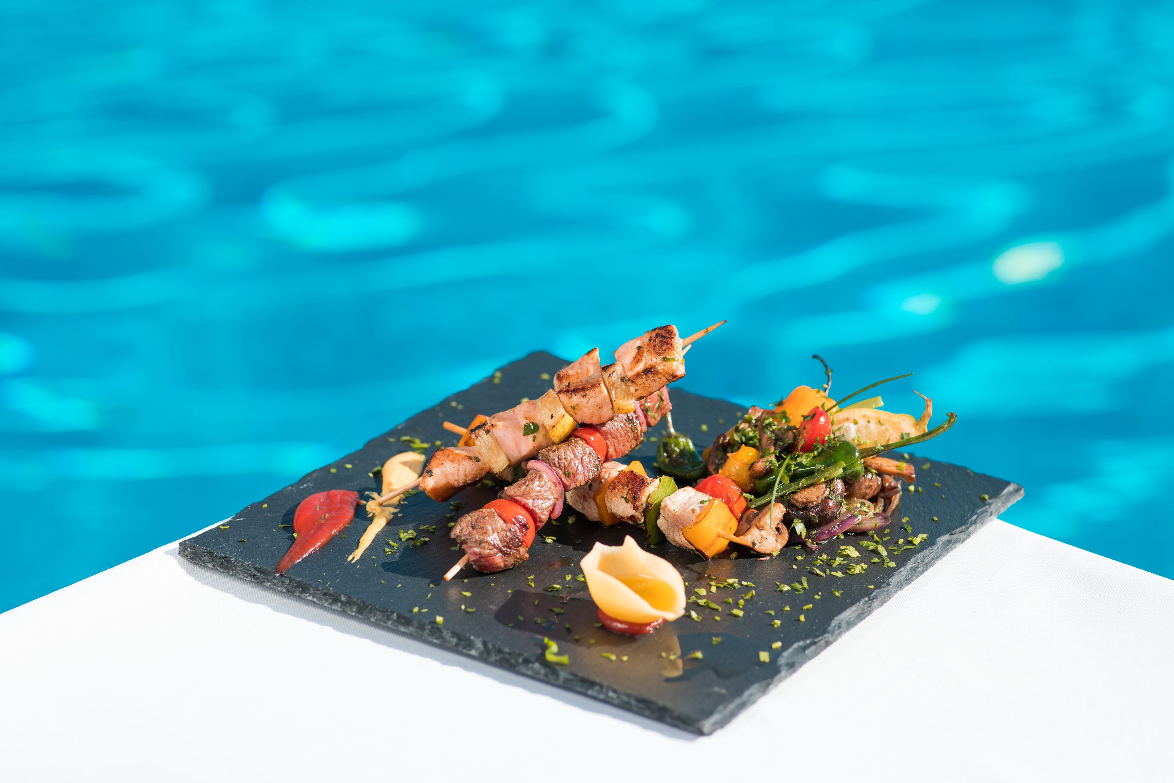 Ein Teller mit Spießen, belegt mit Fleisch und Gemüse, mit einem Pool im Hintergrund.