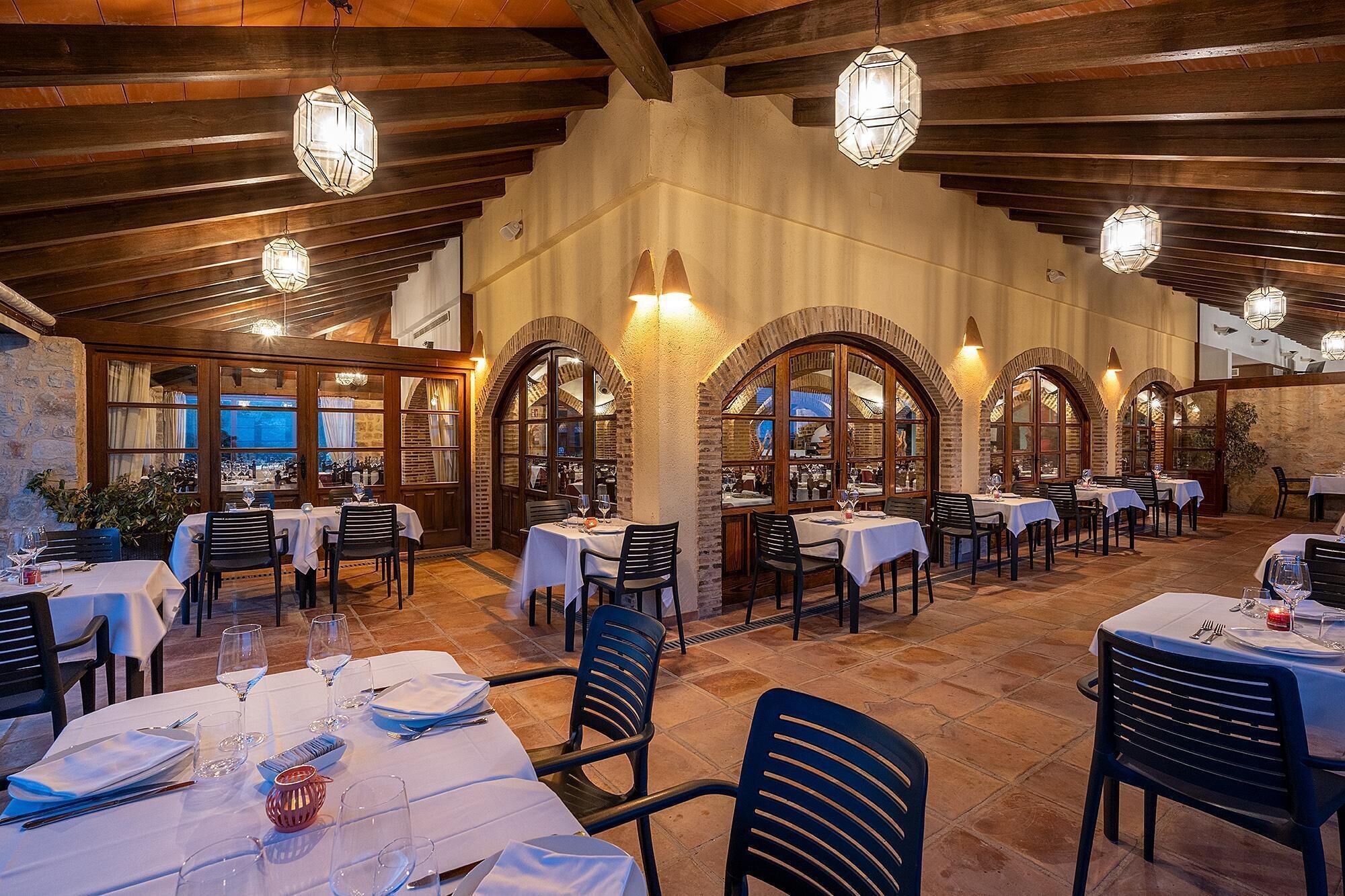 A restaurant with multiple tables and chairs, wooden beams on the ceiling, and arched windows.