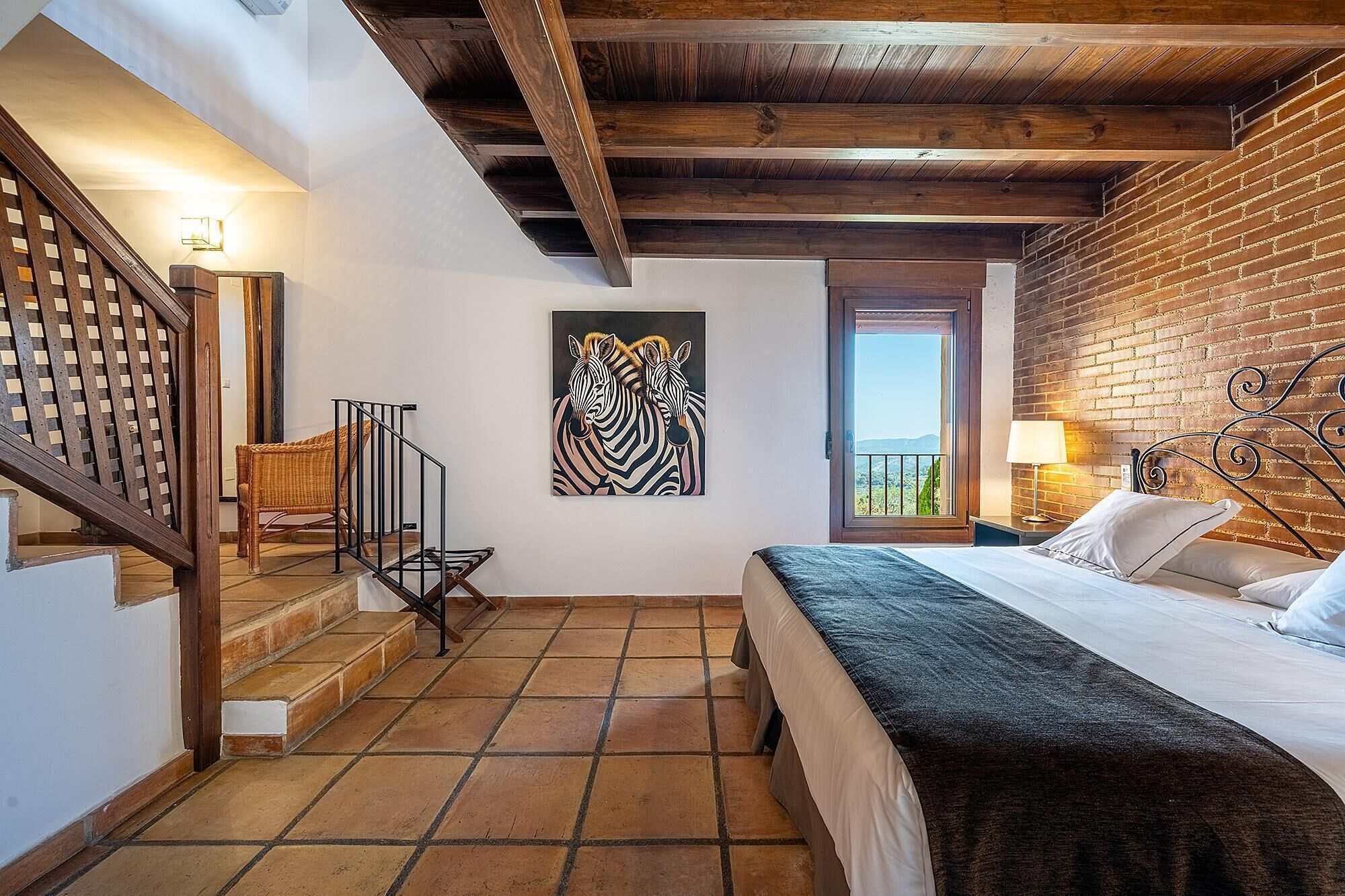 A bedroom with a bed, a window with a view, a staircase, and a zebra painting on the wall.