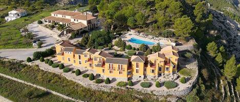 Une grande villa jaune, dotée de nombreux balcons, d’une piscine et entourée d’un paysage rocheux et de verdure.