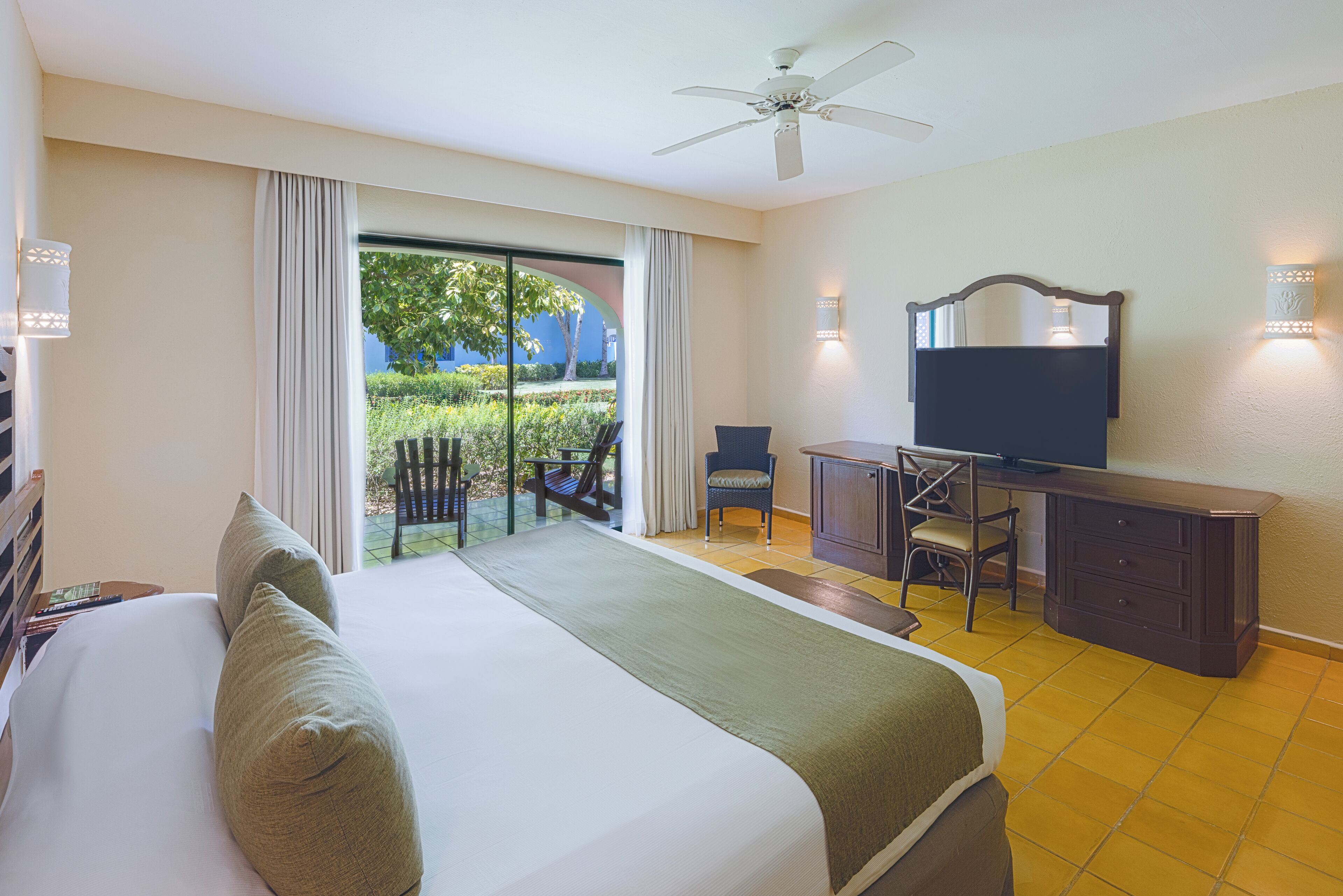 A hotel room with a large bed, a desk with a chair, a television, and a balcony with a view of greenery.