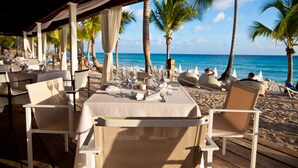 Un espace de restauration en plein air avec des tables pour deux, offrant une vue sur une plage bordée de palmiers et sur l’océan.