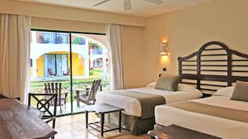 A hotel room with two beds, a desk, and a chair. There is a balcony with a view of a building and a garden.