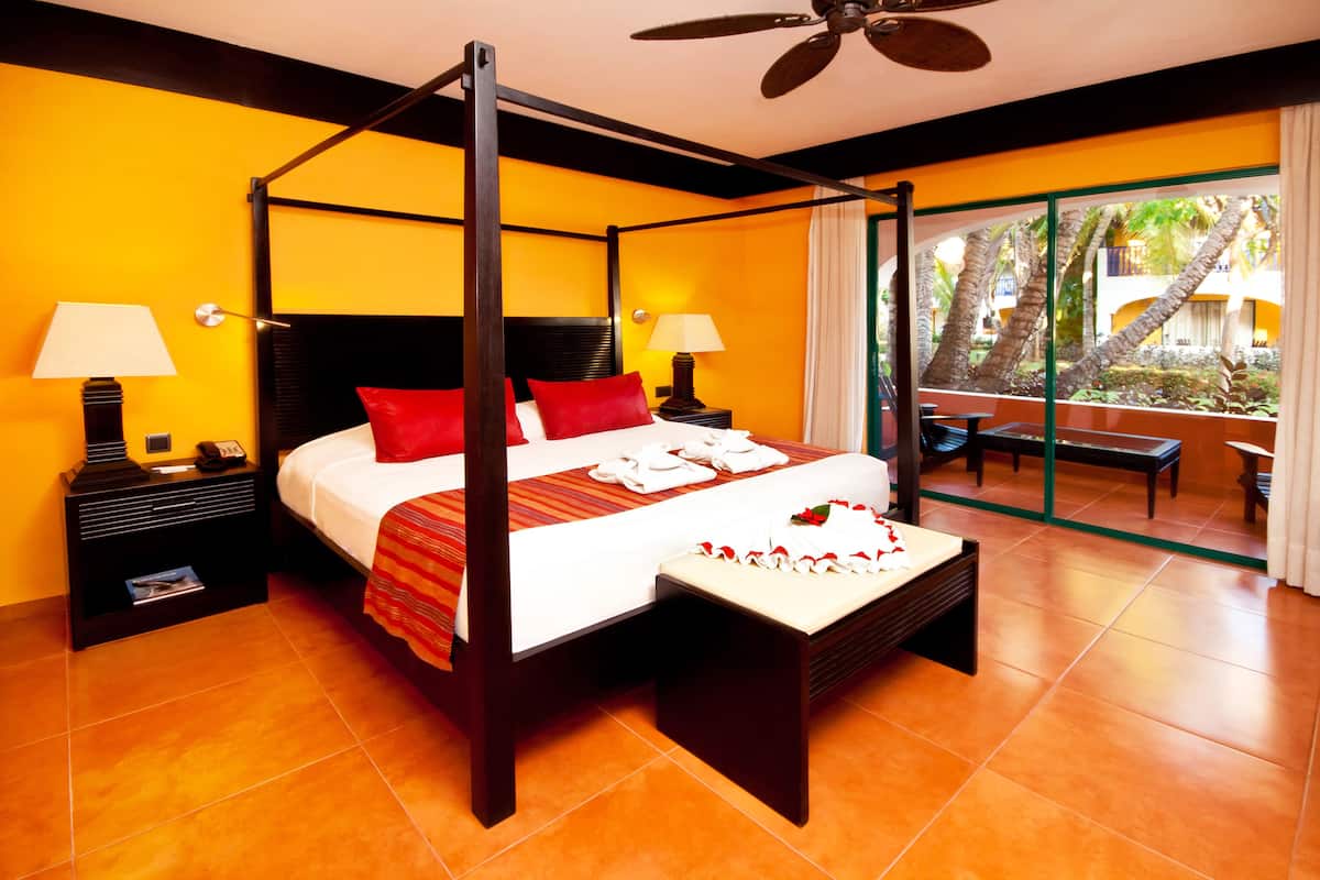 A four-poster bed with red pillows, a bedside table with a lamp, and a view of a palm tree garden.