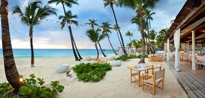 A beachside restaurant with outdoor seating, palm trees, and a view of the ocean. - Catalonia Bayahibe – All Inclusive (San Rafael del Yuma)