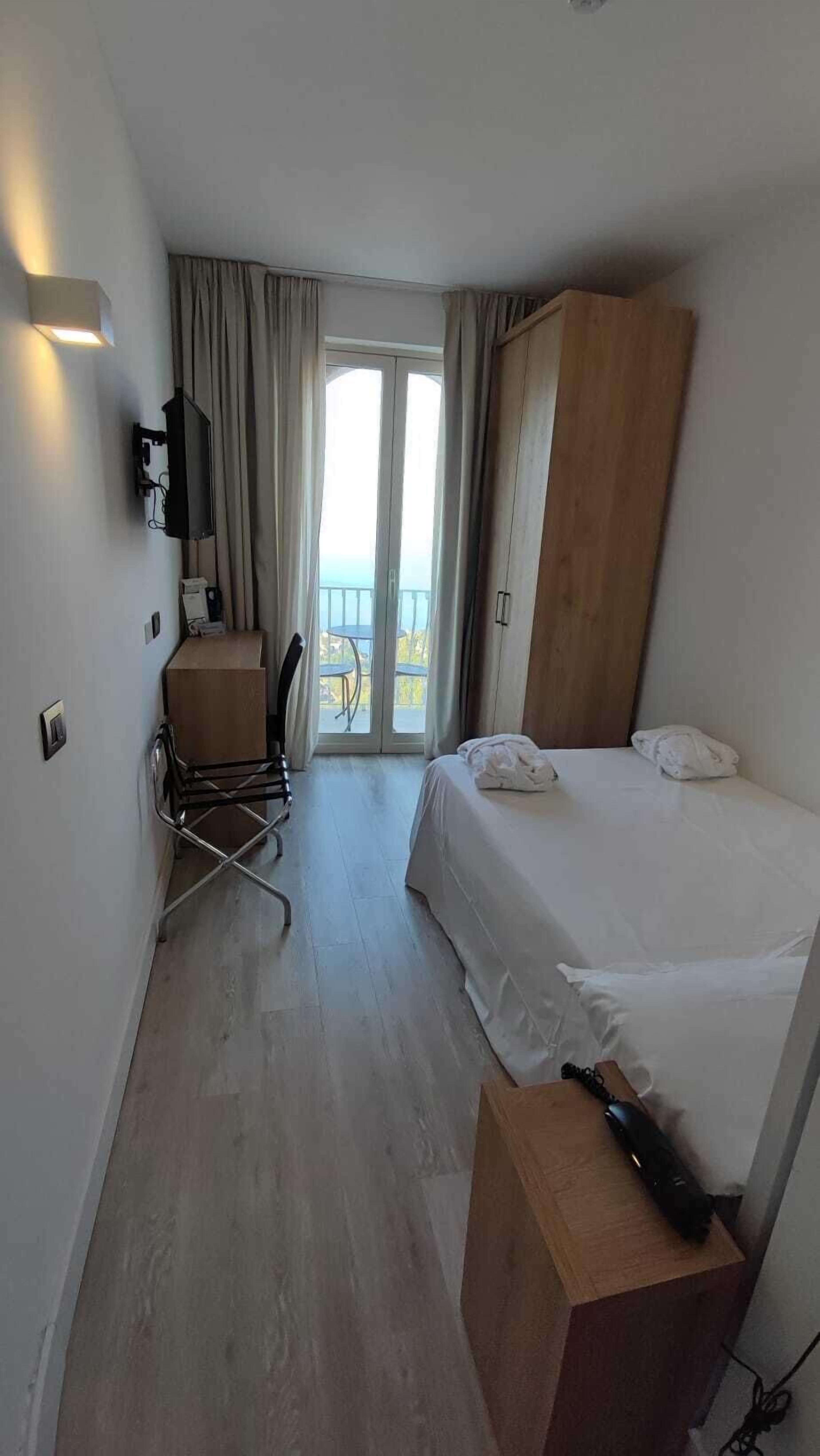 Et hotelværelse med en seng, et skrivebord med stol, et klædeskab og udsigt til det fri gennem en glasdør.