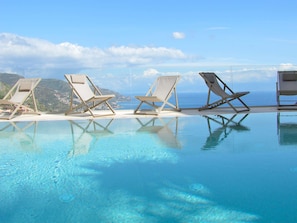 Piscina azul cristalina con sillas de descanso mirando al mar, rodeada por una terraza blanca.