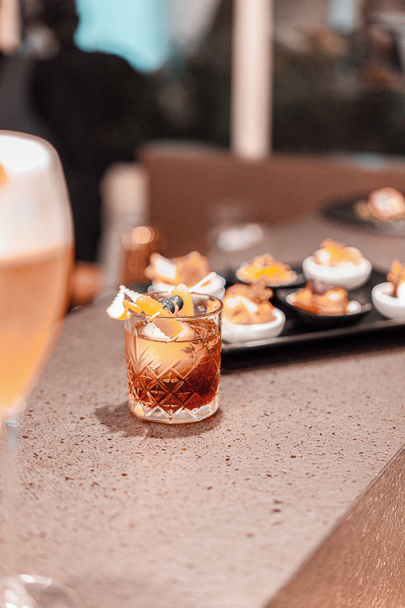 A bar setting with a glass of amber-colored drink garnished with an orange slice and a blueberry, and a tray of small desserts in the background.