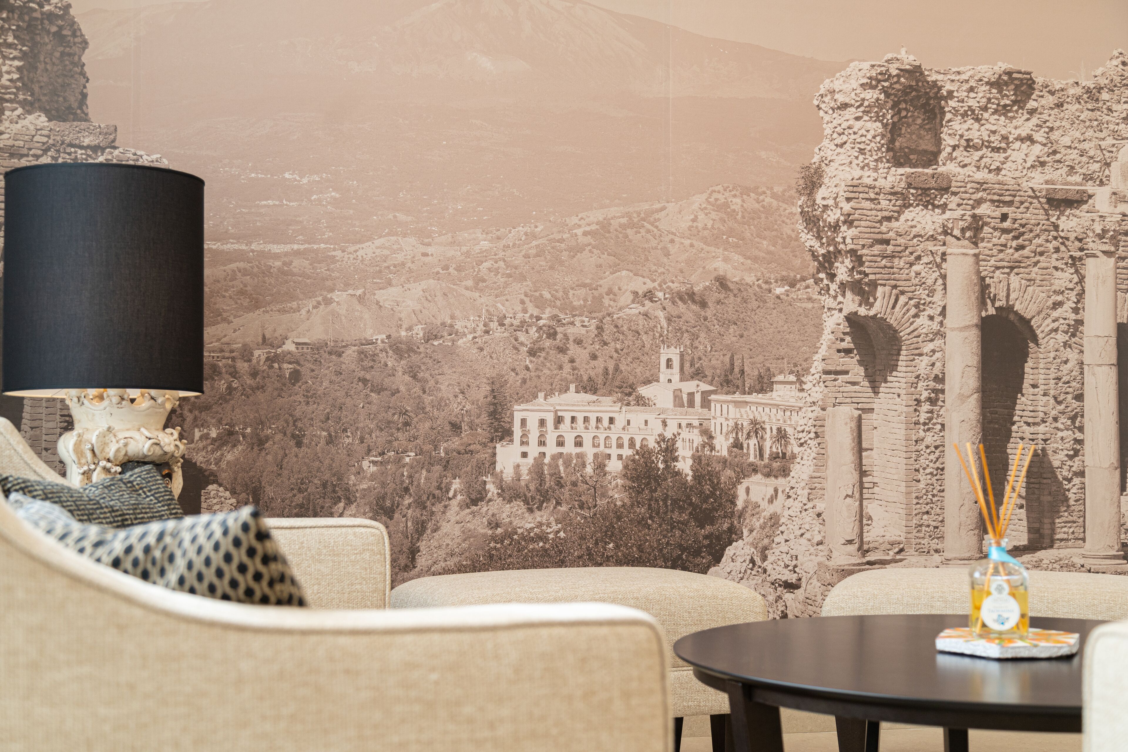 A room with a view of a mountain and an ancient building. There is a lamp, a table with a bottle, and a sofa with patterned cushions.