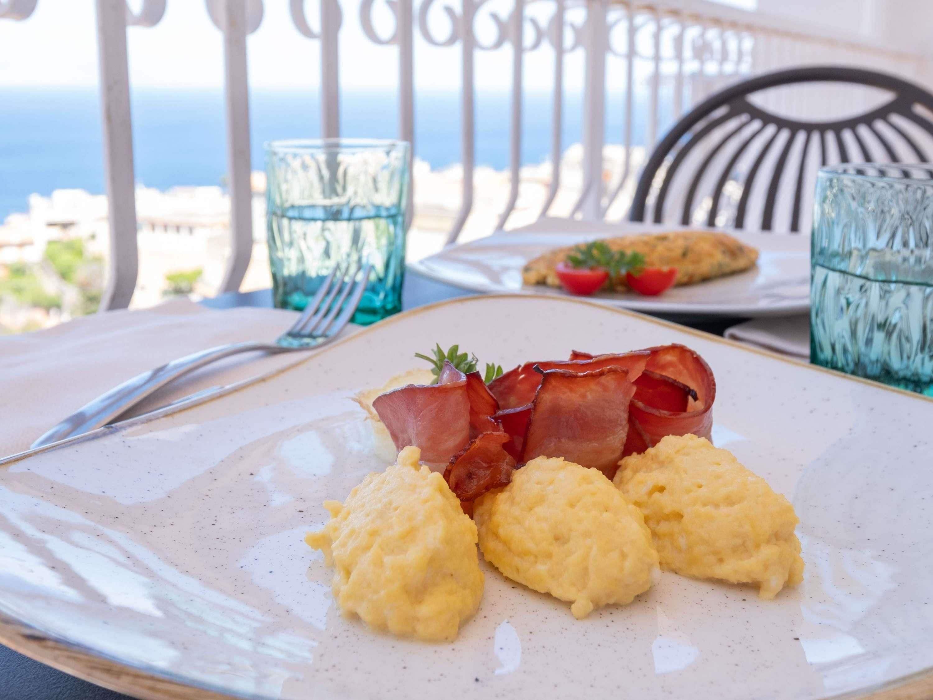 A breakfast plate with scrambled eggs and bacon, a fork, and a glass of water on a table with a view of the ocean.