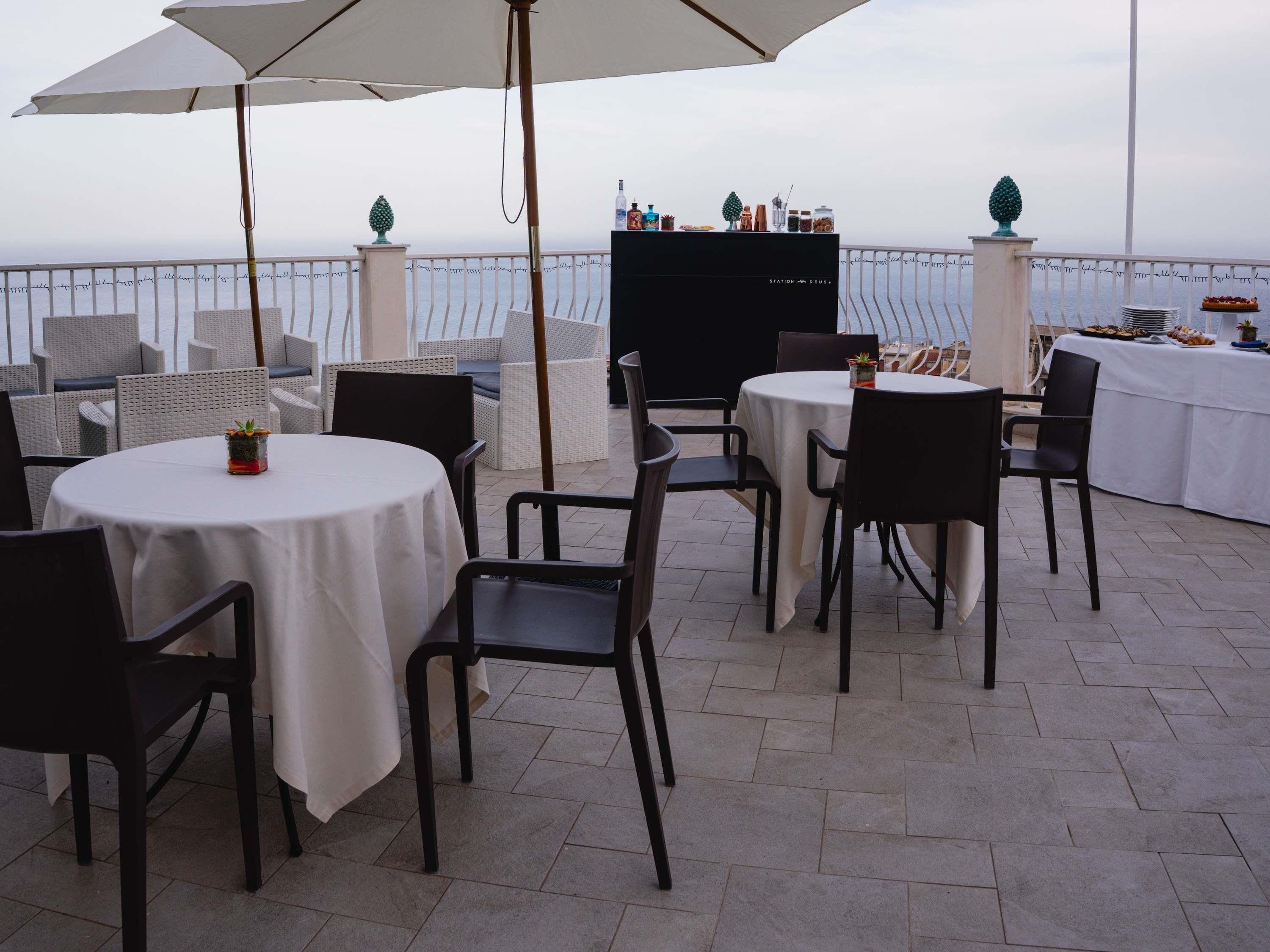Une terrasse extérieure avec des tables et des chaises rondes, un parasol blanc et une vue sur la mer.