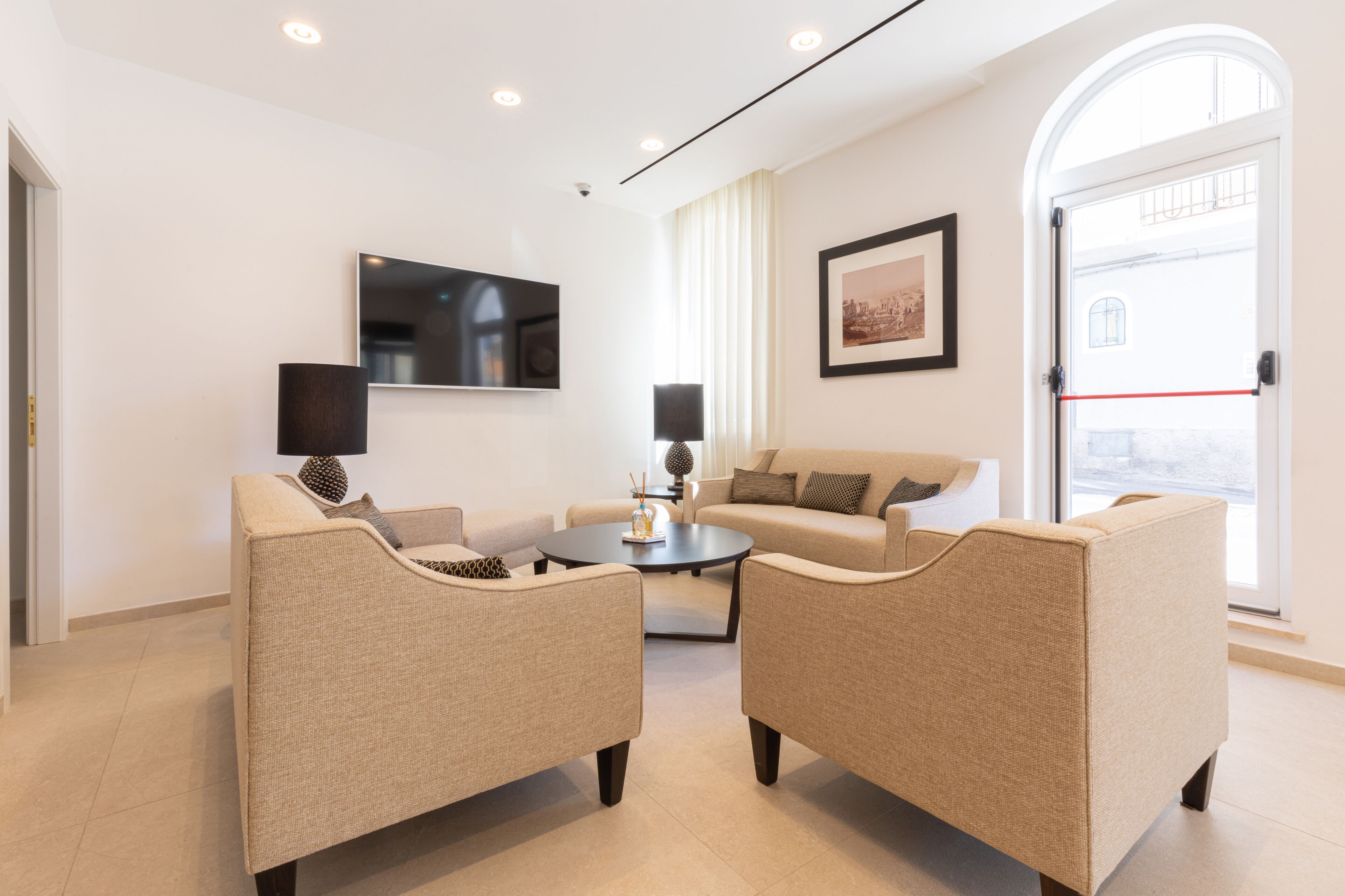 Un salon moderne avec deux fauteuils beiges, une table basse ronde, un canapé, un téléviseur à écran plat et un tableau encadré accroché au mur.