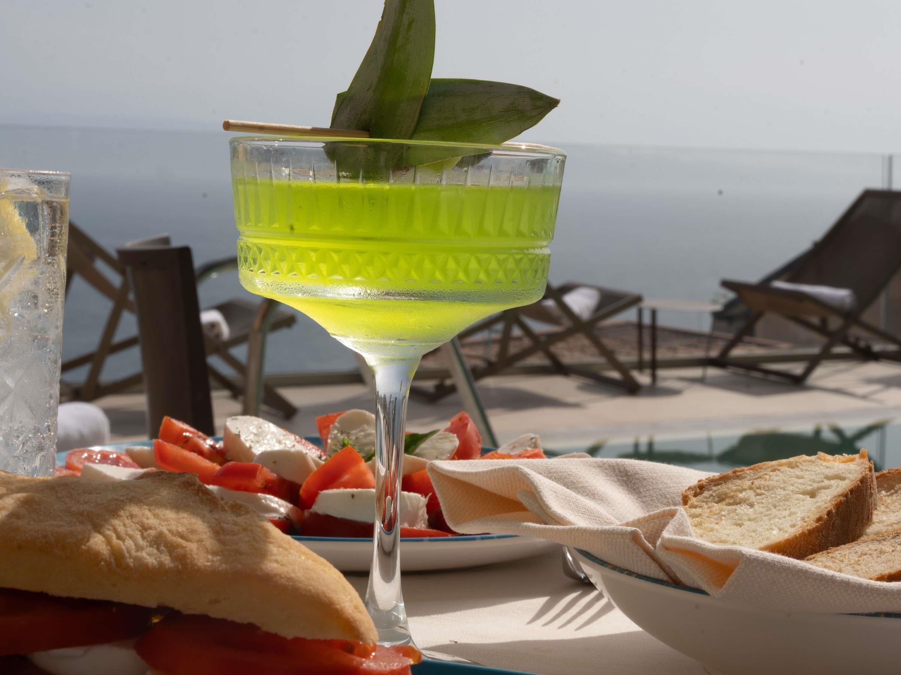 A table with a glass of a yellow beverage, a plate of food, and a view of the sea.