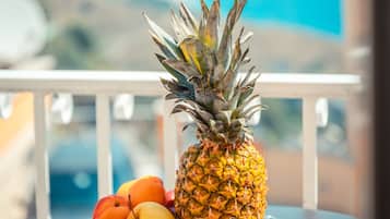 Una mesa con una cesta de frutas que contiene una piña, una manzana, una pera y un kiwi, en un balcón con vista al océano.