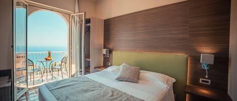 Habitación de hotel con cama, mesita de noche, lámpara de pared, vista al mar y balcón con sillas y una mesa.