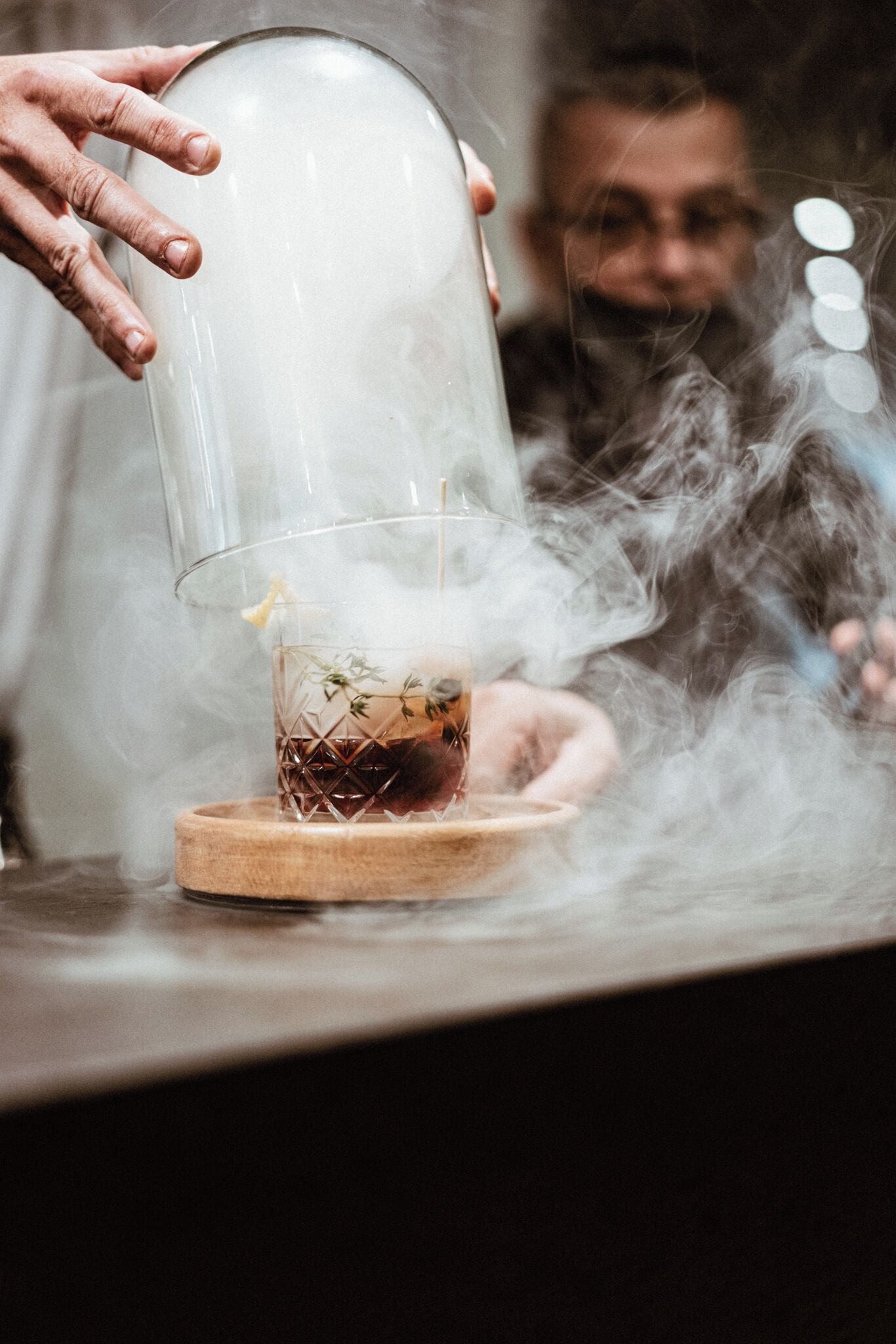 A person pouring a drink from a glass dome into a glass on a wooden coaster.