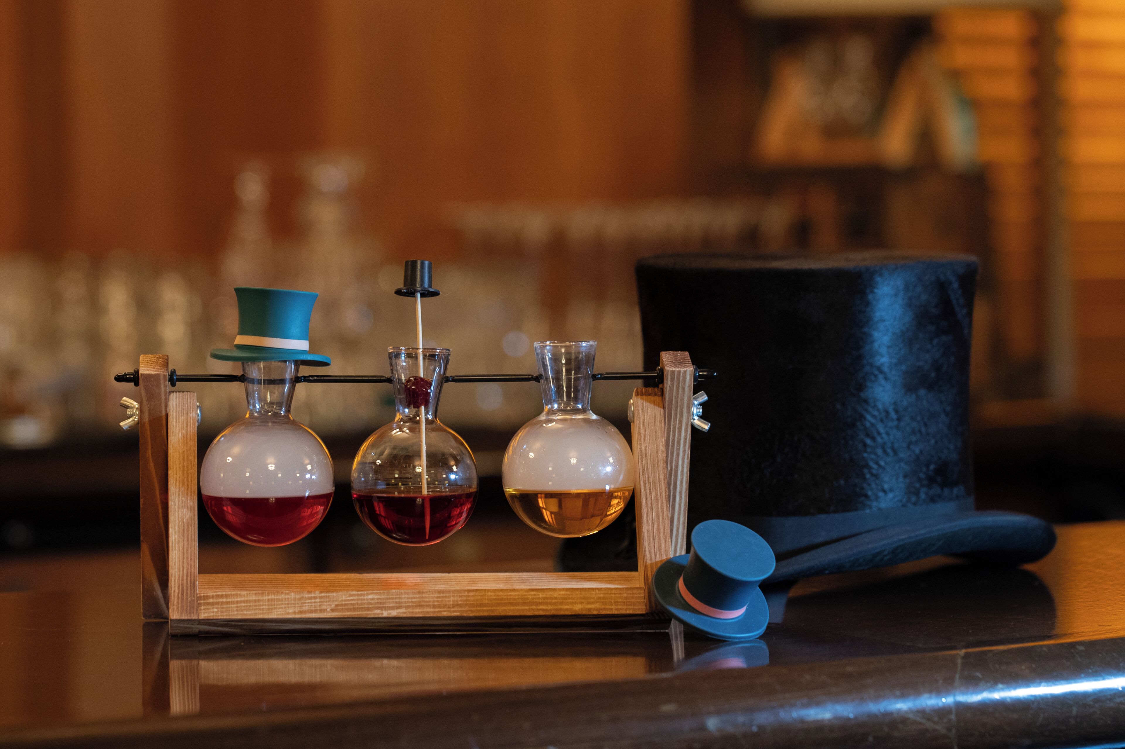 A wooden apparatus with three glass containers, each holding a different colored liquid, and a black top hat beside it.