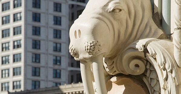 Primer plano de una escultura arquitectónica blanca con delicados grabados, frente a un edificio moderno con múltiples ventanales.